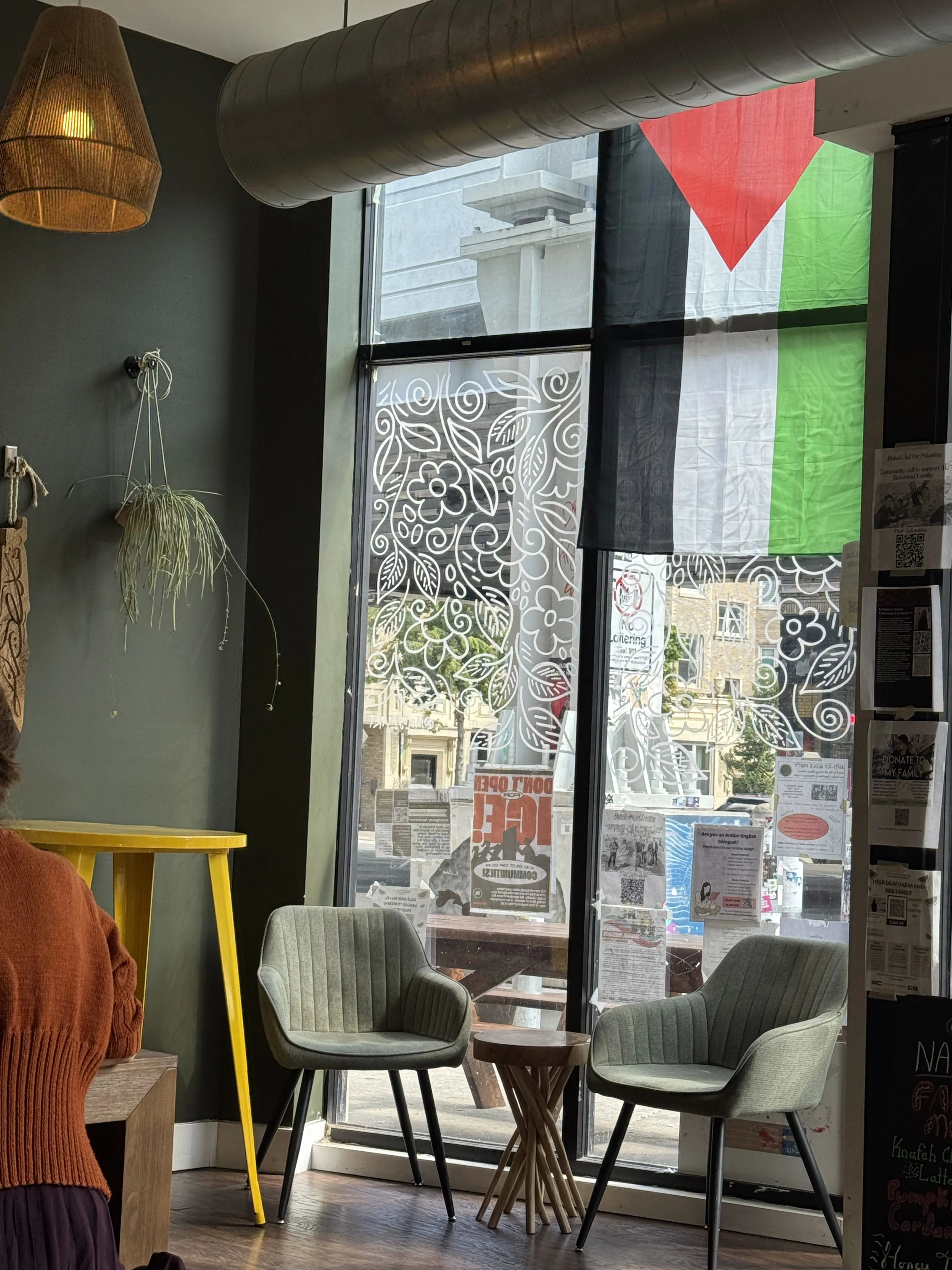 Front of Nabala cafe, focus on two sage green chairs and the large window with olive branch decals and a Palestinian flag