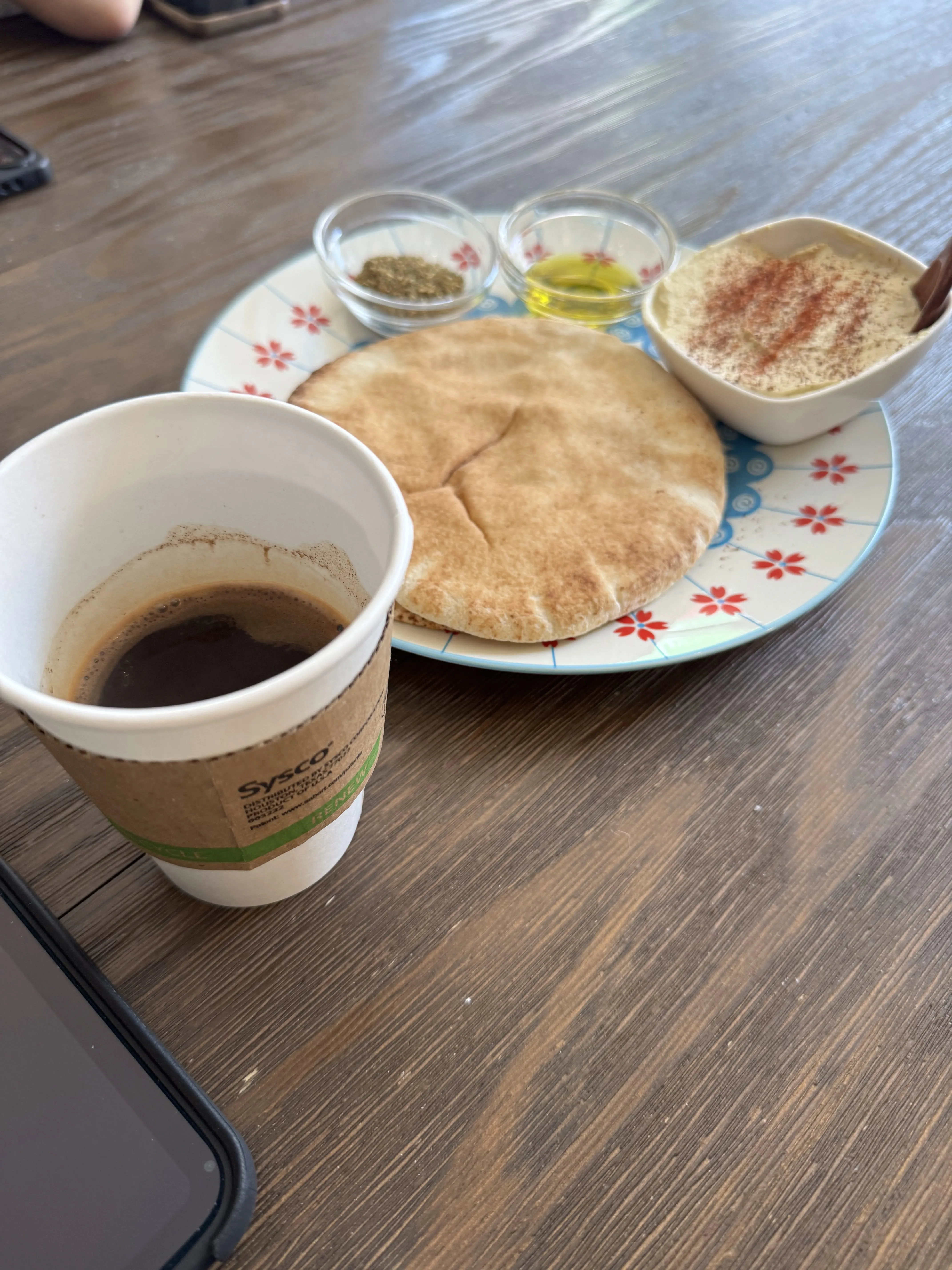 Turkish coffee in a to-go cup next to a nice plate with 2 loaves of pita bread, hummus, za'atar mix, and olive oil