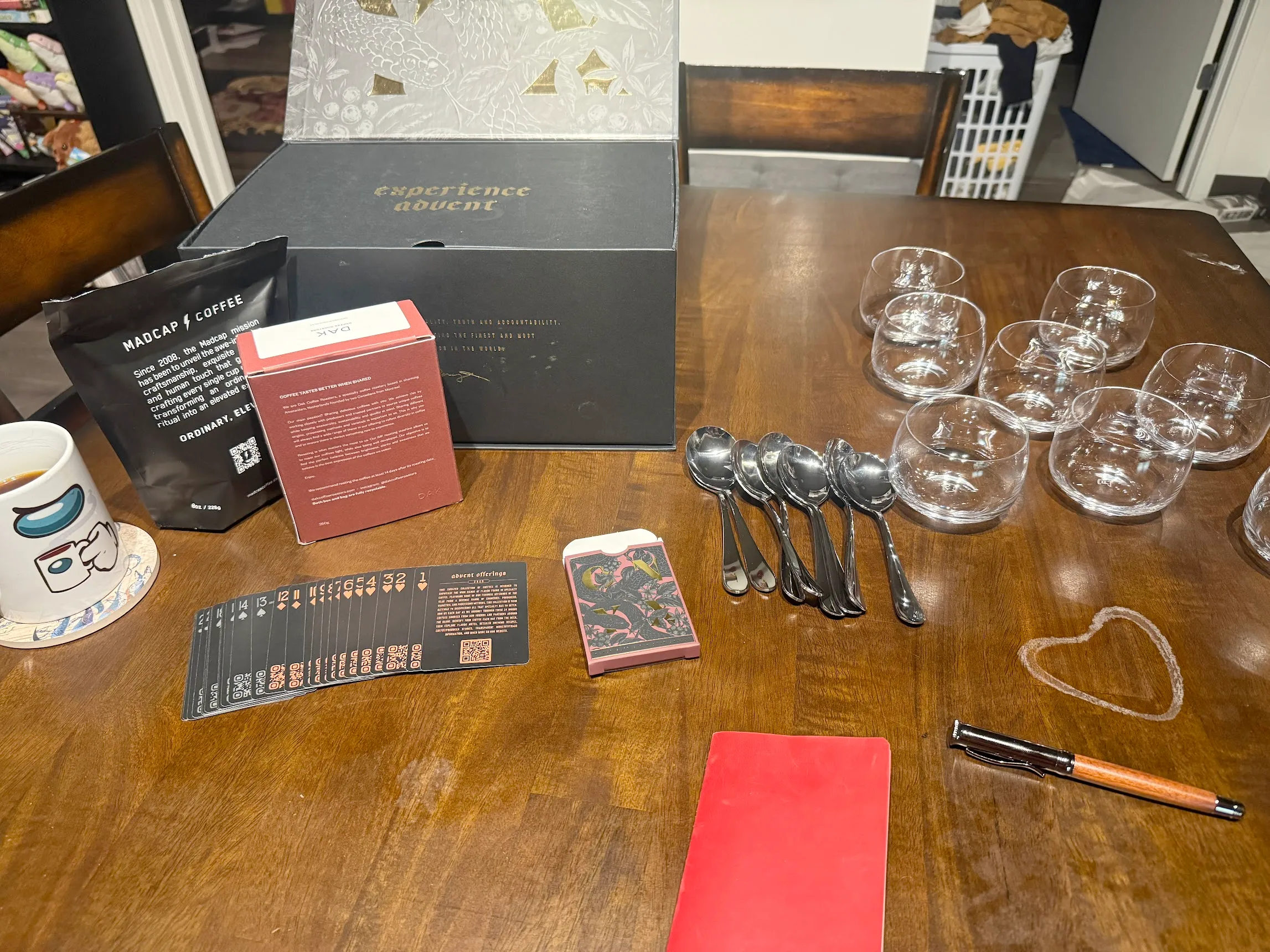 Pre-cupping setup. The Onyx coffee advent box is in the back. There are a bag and box of coffee in front of it, next to an amongus coffee mug (amug-us). Next to that is a spread out mini-deck of cards with coffee info written on them. To the right side are 8 shallow glass cups next to 8 spoons, a small red notebook, and pen.