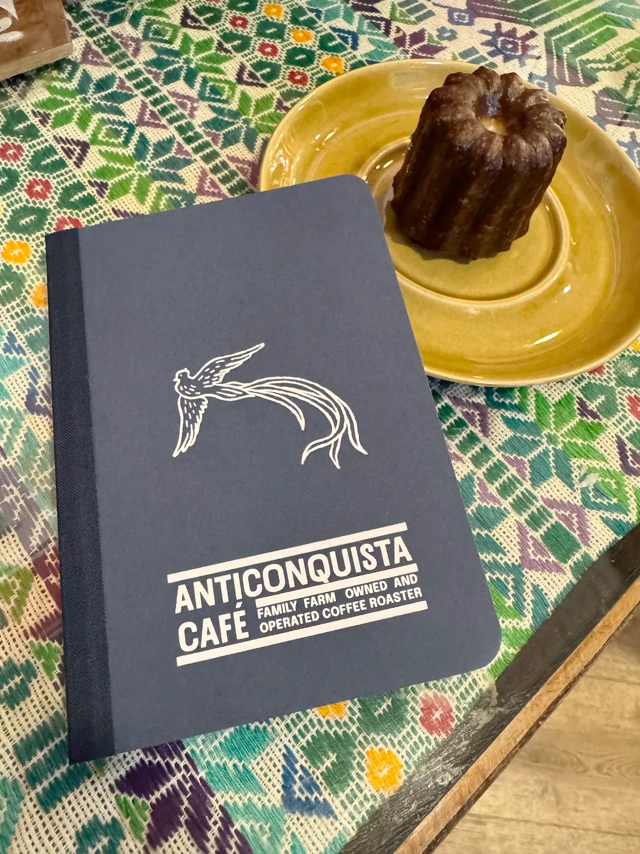 crazy how well this low-effort picture turned out A blue notebook labeled Anticonquista Cafe Family Farm Owned and Operated Coffee Roaster atop a green and blue woven mat beside a yellow plate with a brown cylindrical pastry