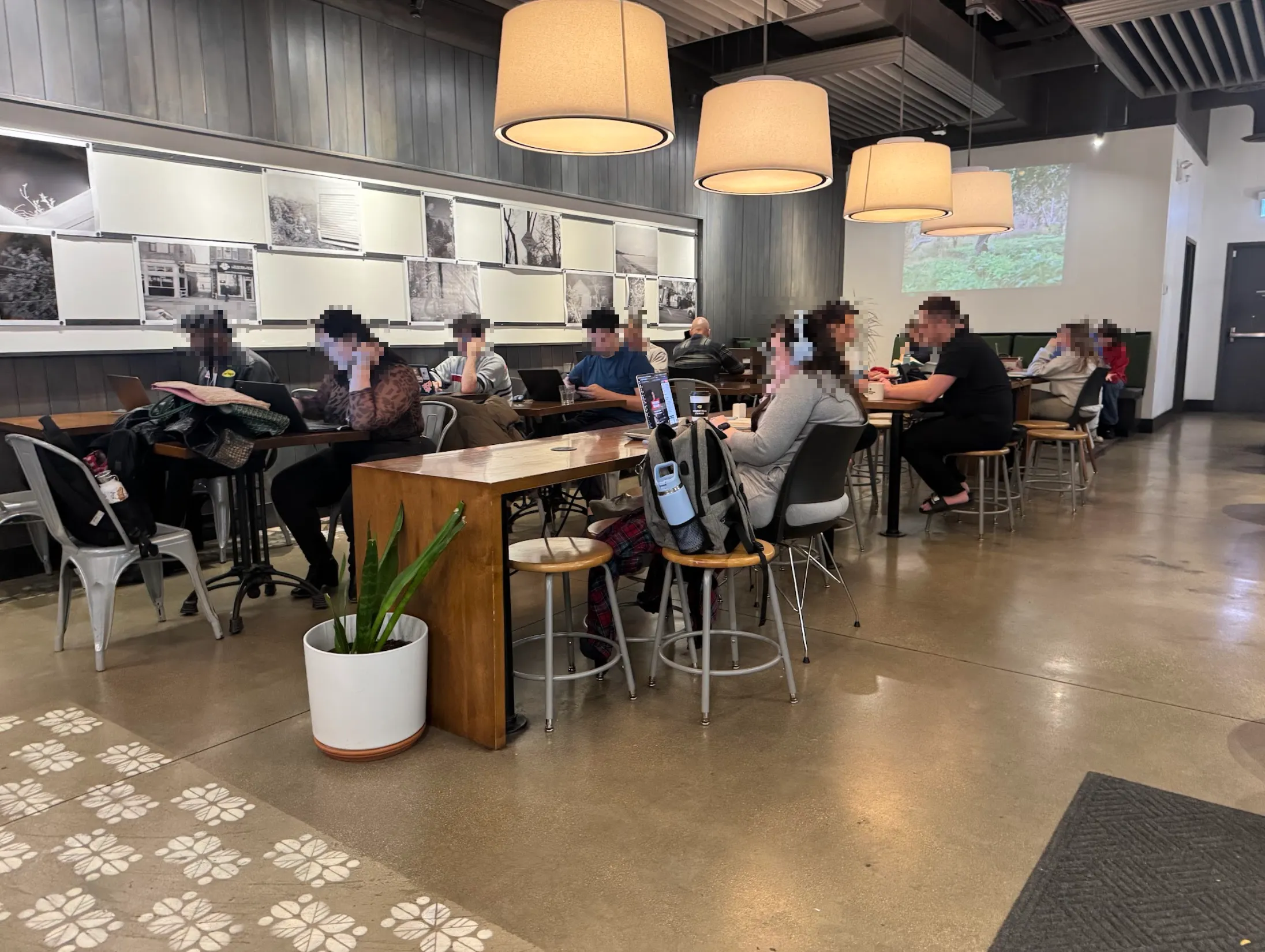 Large, standard cafe space with a few long bar-style tables in the center, and 2 columns of cafe 2-person tables behind. There are many people here, all with faces blurred out.